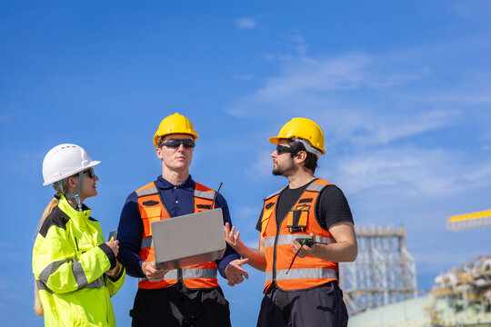 LNG plant engineers team discussing work with laptop at terminal, liquefied natural gas workers meeting at industrial facility, energy industry professionals talking together outdoors under blue sky