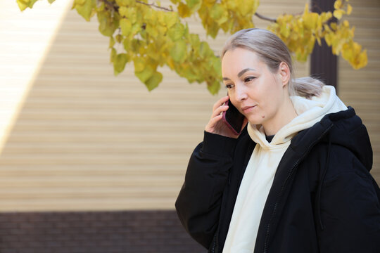 Young Woman In Black Coat And White Hoodie Talks On Smartphone Outdoors Beside Soft Yellow Autumn Leaves And Sunlit Building Facade, Creating Calm Urban Lifestyle Scene 