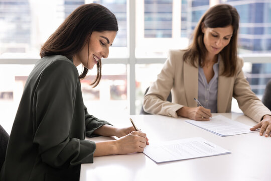 Two satisfied diverse female professionals business partners signing agreement copies at meeting table approving collaboration establishing partnership confirming mutual decision in official contract