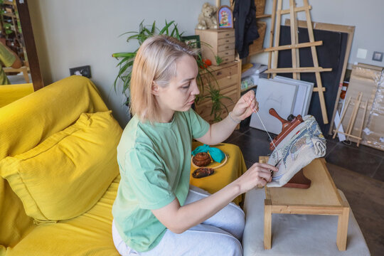 Young Woman In Mint Green Shirt Carefully Embroidering Fabric On Hoop Beside Bright Yellow Sofa Inside Cozy Home Studio, Surrounded By Plants, Easels, Drawers, Framed Art, And Crafting