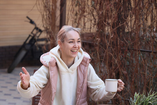 Young Woman Smiles While Holding White Mug And Gesturing During Relaxed Conversation In Outdoor Garden Patio, Wearing Cozy Hoodie And Pink Vest Beside Dry Vines, With Soft Natural Light