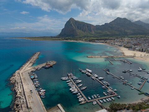 Aerial view of the marina in San Vito Lo Capo with numerous boats docked in turquoise water, a sandy beach, and the prominent Monte Monaco mountain in the background in Sicily, Italy.