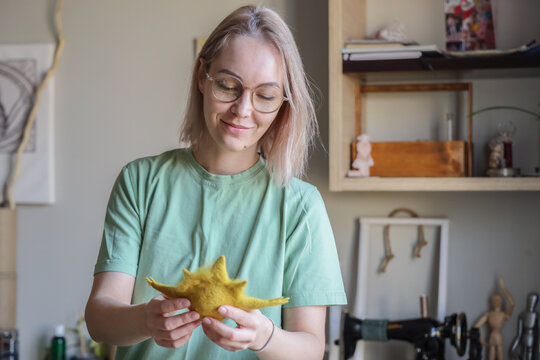 Young Woman With Glasses Carefully Holds Soft Yellow Felt Figure In Cozy Home Studio, Showing Handmade Textile Work, Crafting Hobby, Sewing Tools, Shelves