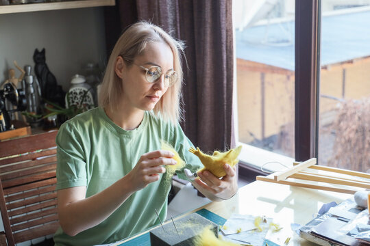 Blonde Woman With Glasses Works On Felt Animal Figure At Sunlit Home Workspace, Holding Soft Wool While Shaping Details With Needle Felting Tools And Focused Attention 