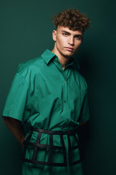 Young man with curly hair wearing green shirt and black striped harness standing against green background in studio. Fashion portrait and casual pose with serious facial expression and male model.