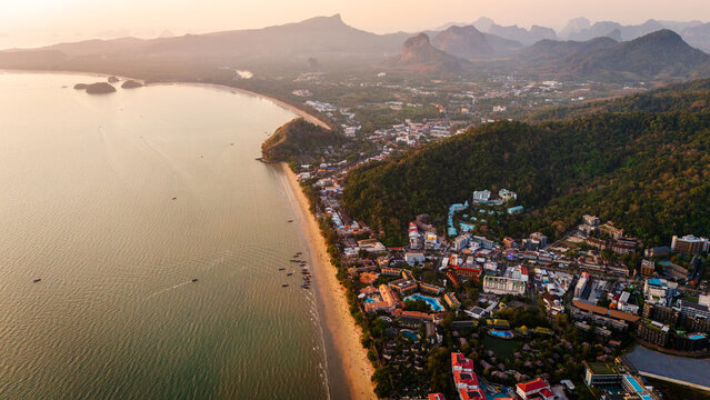 Aerial view of the coastline of Ao Nang featuring sandy beaches, lush green hills, hotels, and boats on the water at sunset in Krabi, Thailand.