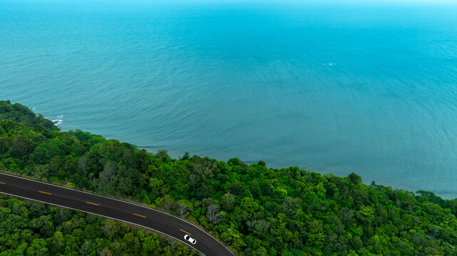 Top view aerial shot of a white car driving on a coastal road between lush green forest and turquoise ocean water, concept of scenic nature landscape representing travel and exploration.	