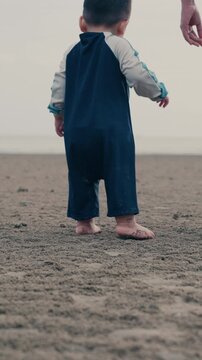 Back view of cute Asian toddler taking first steps walking barefoot on sandy beach.