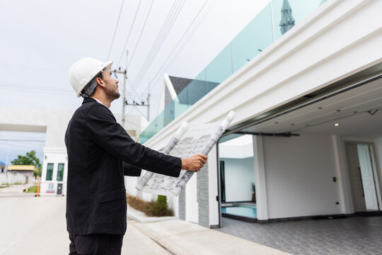 Professional male engineer wearing a safety helmet and holding an architectural blueprint while inspecting the progress of a residential construction project. 