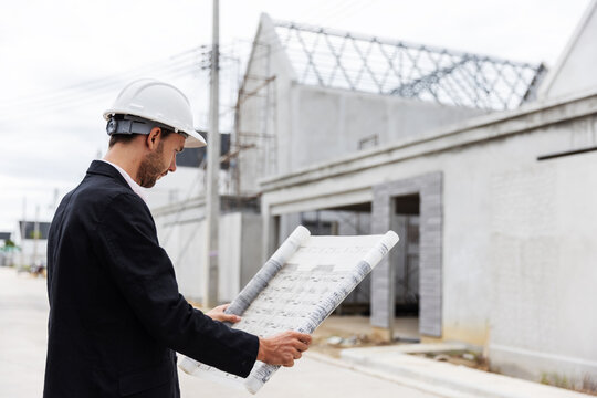 Professional male engineer wearing a safety helmet and holding an architectural blueprint while inspecting the progress of a residential construction project. 