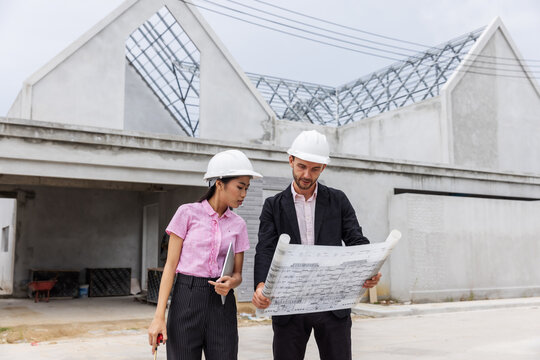 Male engineer and female architect wearing safety helmets while carefully reviewing an architectural blueprint at a residential construction site.
