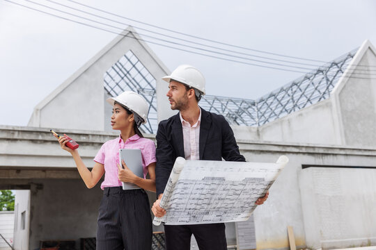 Female architect using a two-way radio and digital tablet to coordinate work while a male engineer reviews a blueprint at a residential construction site. 