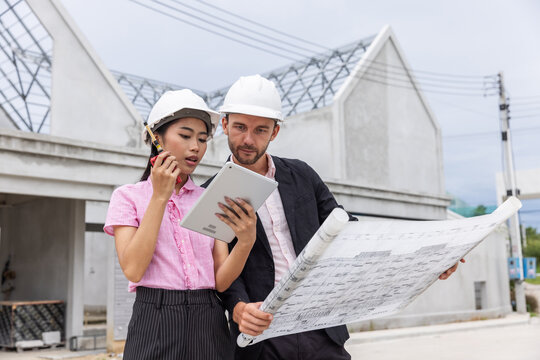 Female architect using a two-way radio and digital tablet to coordinate work while a male engineer reviews a blueprint at a residential construction site. 