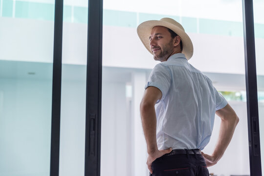 Smiling young man wearing a straw sun hat and standing beside large glass doors in a bright modern residence. Summer lifestyle, vacation mood,