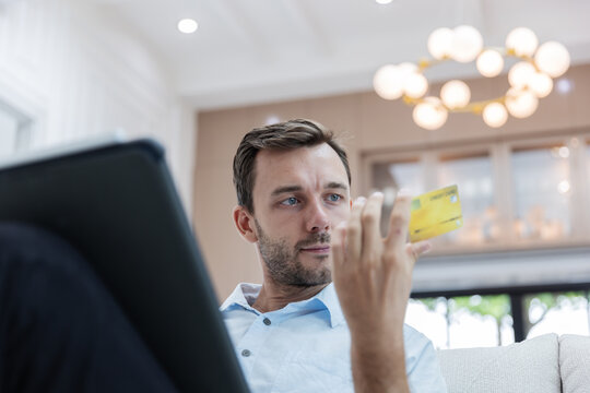 Young man holding a credit card while using a laptop to make an online purchase from home. E-commerce checkout, secure digital payment, internet banking, cashless transaction and consumer technology .