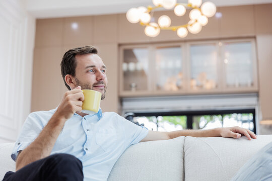 Content young man enjoying a peaceful cup of coffee while relaxing on a comfortable sofa in a bright modern living room. Mindfulness, slow living, morning routine.