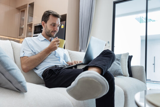 Young professional using a laptop and drinking coffee while relaxing on a sofa in a modern living room. Remote work