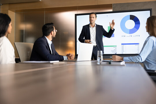 Businessman, corporate trainer presenting financial data displayed on screen with charts and diagrams, share sales statistics, make business presentation for clients, colleagues in office meeting room