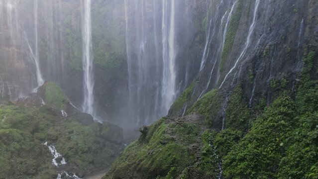 Aerial view of Tumpak Sewu Waterfall featuring cascading water streams over lush green cliffs and rocky terrain in Lumajang, East Java, Indonesia.