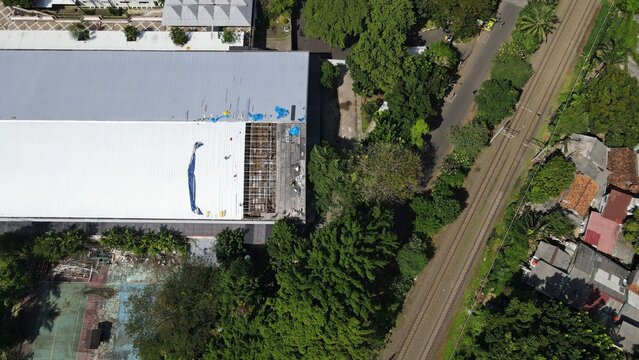 Aerial view of Pertamina Arena, Simprug showing roof repair work with blue tarps and exposed structural elements next to railway tracks in Kecamatan Kebayoran Lama, Daerah Khusus Ibukota, Indonesia.