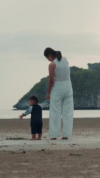 Vertical shot of Asian mother holding hands with toddler son standing on beach.