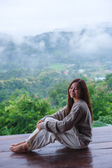 Fototapeta premium Vertical image of a woman sitting on wooden balcony with a beautiful mountain views on cloudy morning