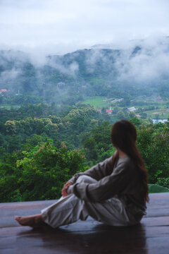 Vertical rear view image of a woman sitting on wooden balcony with a beautiful mountain views on cloudy morning