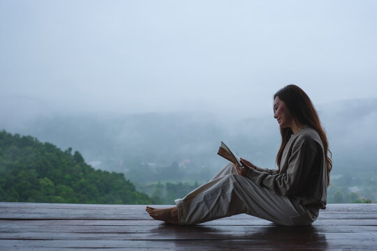 A woman reading a book while sitting on wooden balcony with a beautiful mountain views on cloudy morning