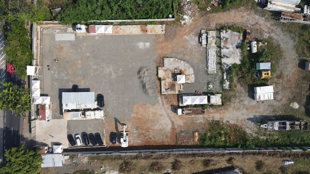 Aerial view of Bambu Apus construction site with gravel ground, parked vehicles, white tents, and machinery in a fenced area in Kecamatan Cipayung, Daerah Khusus Ibukota, Indonesia.