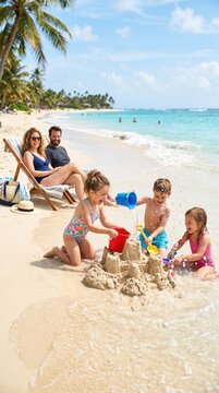 Family beach vacation showing parents sitting on lounge chair while three children build sandcastle and play with toys at ocean shore.