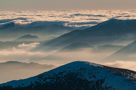 Aerial view of mountain horizons as seen from Dumbier with snow-capped peaks rising above a sea of clouds and sun rays filtering through the mist in Nizke Tatry, Zilina Region, Slovakia.
