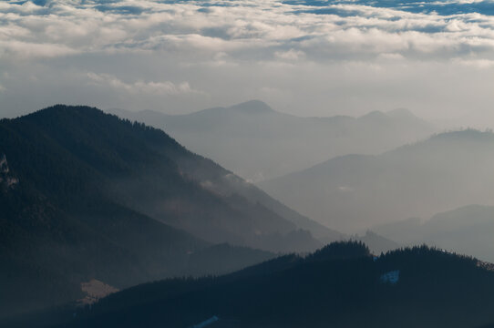 Aerial view of the mountain horizons as seen from Dumbier, featuring layered peaks, misty valleys, and dense pine forests under a cloudy sky in Nizke Tatry, Zilina Region, Slovakia.
