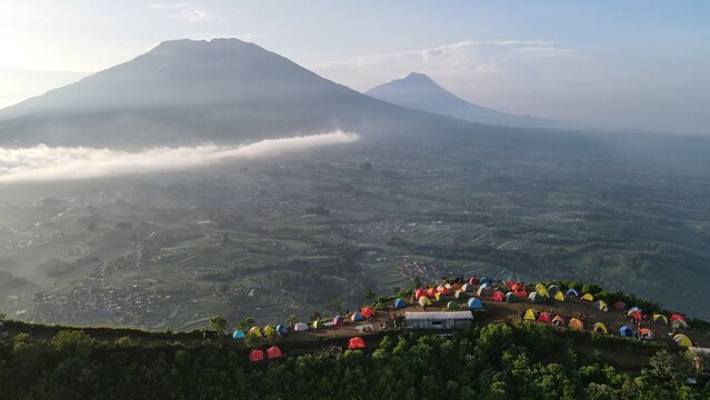 Aerial view of Gunung Andong Via Pendem with colorful camping tents on the ridge, surrounded by lush terraced fields and morning mist in Kecamatan Ngablak, Jawa Tengah, Indonesia.