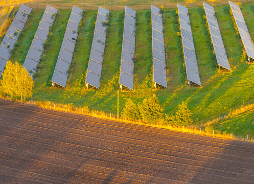 Aerial view of solar panels arranged in rows on a green hillside next to a freshly plowed brown field under warm golden hour light in Gniew, Pomeranian Voivodeship, Poland.