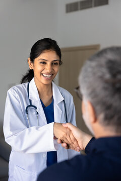Smiling South Asian female doctor in white coat shaking hand, greeting male patient in clinic office. Trust in healthcare services, professional consultation, positive outcome of medical visit concept