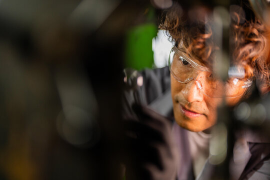 Mature Asian technician at his traditional machining workshop, dad's face right-of-center peers down-left between out-of-focus machine parts, skilled work of a family enterprise founder