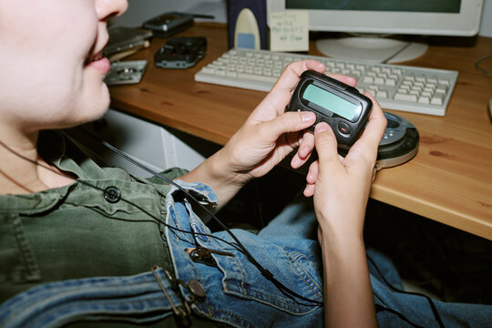 Young adult Caucasian woman holding retro pager at office desk with vintage computer, communicating in nineties style. Image suitable for nostalgic tech marketing campaigns