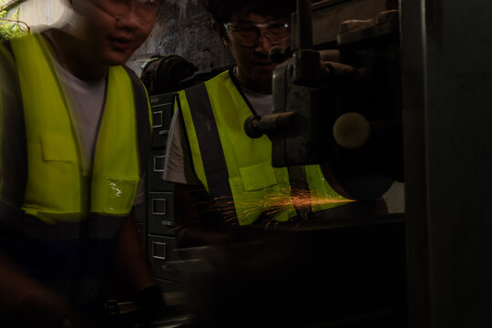 Asian father and teenage son at the family machining workshop, son motion-blurred in left foreground, dad sharper at right, a small of sparks, father teaching son the trade to inherit