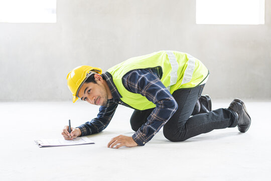 Asian male construction engineer or inspector in safety uniform checking concrete floor quality and writing on clipboard checklist at home building renovation site project.