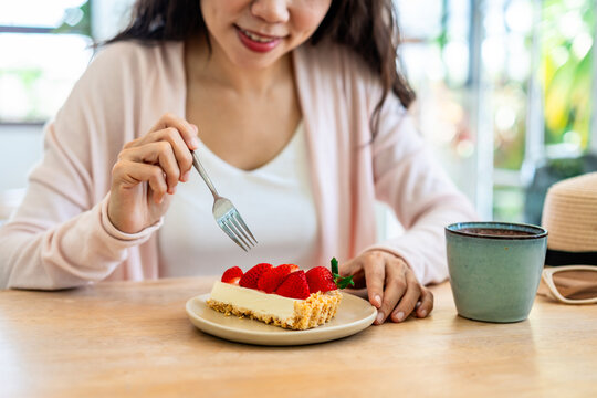 Cropped shot of a happy young woman holding a slice of strawberry cake and a fork, enjoying dessert in a cozy cafe