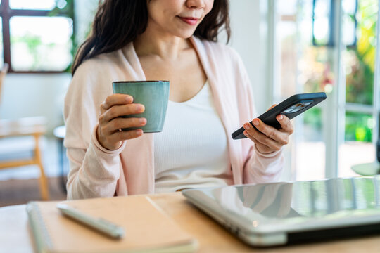 Young woman holding a coffee cup and checking her smartphone at a desk with a laptop and notebook; digital nomad and remote working concept