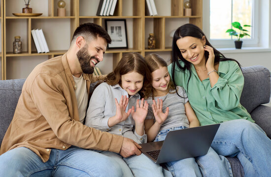 Family parents children sisters join laptop online videocall. Parents with two daughters on a sofa at home, laughing and waving to friends online. Concept conveys joyful digital togetherness