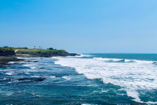 Eye level landscape photograph of rolling sea waves, white foam, and green coastal cliffs under clear sky at Tanah Lot, Bali, Indonesia. Perfect for travel , banner ads, and nature themes.