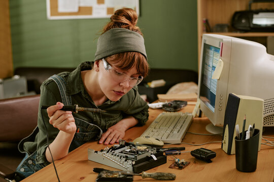 Young adult Caucasian woman wearing safety glasses soldering computer hardware in retro office. Image represents vintage technology repair and is suitable for Information Technology service marketing