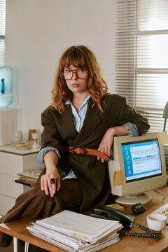 Portrait of Caucasian young adult woman sitting on desk in retro office next to vintage computer monitor, representing corporate nostalgia and 1990s workplace culture, vertical