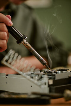 Vertical image of hand of Caucasian young adult holding soldering iron over vintage electronic device with smoke rising, representing retro office repair and engineering concepts