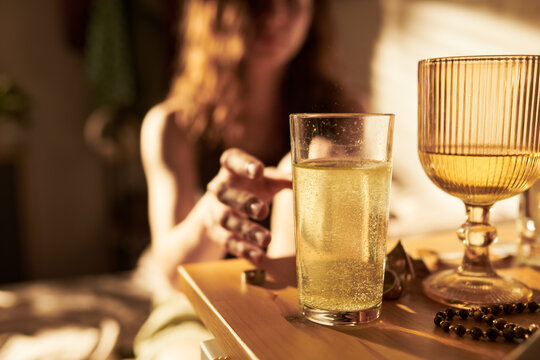 Caucasian young woman reaching for glass of effervescent yellow liquid on table, representing morning hydration or hangover cure. Image suitable for wellness and dietary supplement marketing