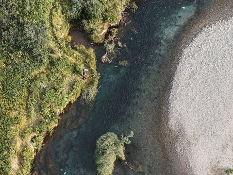 Aerial view of a fisherman standing on the grassy bank of a clear river with a gravel bed in Unalaska, Alaska, United States.