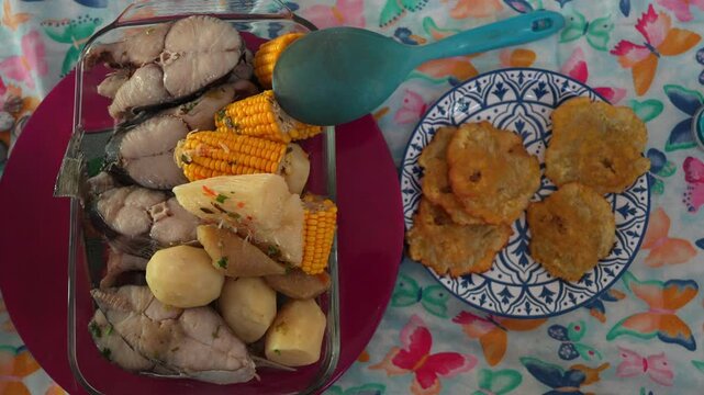 Overhead shot of freshly prepared Colombian sancocho with fish, potatoes, corn, and yuca beside golden patacones on patterned plates atop a colorful floral tablecloth.