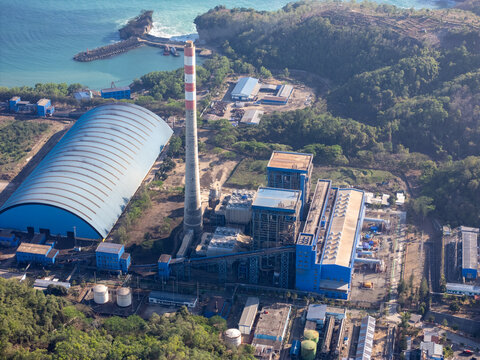 Aerial view of the Pacitan Power Plant with a tall chimney, blue storage buildings, and industrial facilities along the coast near green hills in Pacitan, East Java, Indonesia.